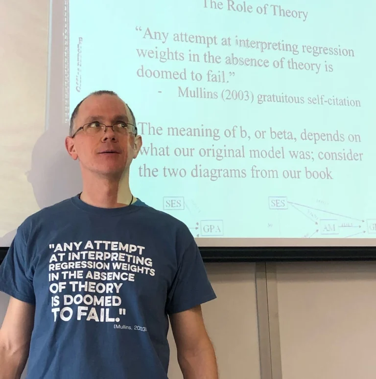 Morrie, wearing a shirt that reads, "Any attempt at interpreting regression weights in the absence of theory is doomed to fail," citing himself. He stands in front of a screen on which the same quote is projected, citing himself. Picture is from a graduate statistics class.
